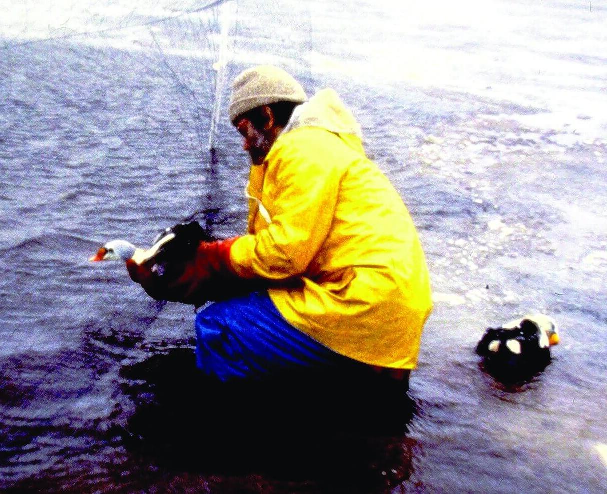 一名戴着无边帽、穿着黄色雨衣的志愿者蹲在含油的水中，用戴着手套的手从网中捞出一只深色身体、头部为白色和橙色的鸟.